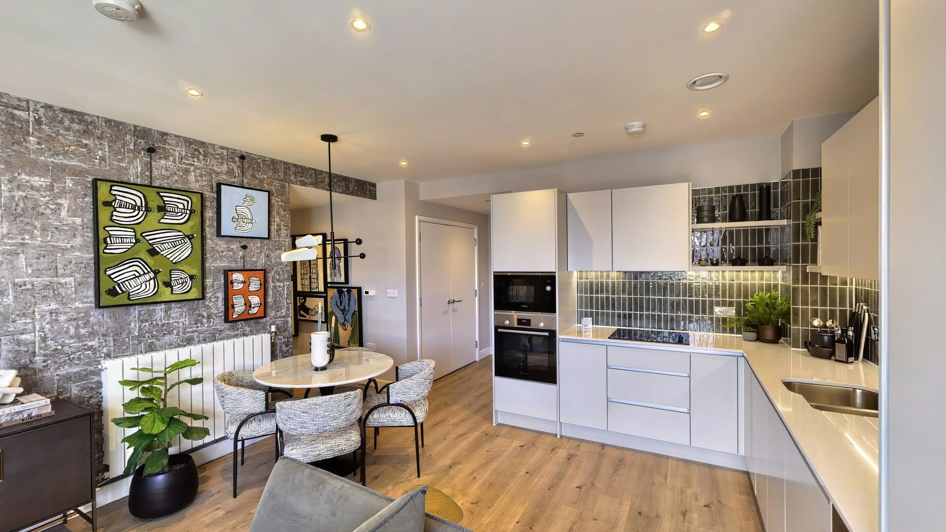 Dining area in the Fero House show apartment: a round marble table beneath a textured metallic feature wall hung with bold bird prints, with a green-tiled kitchen beyond