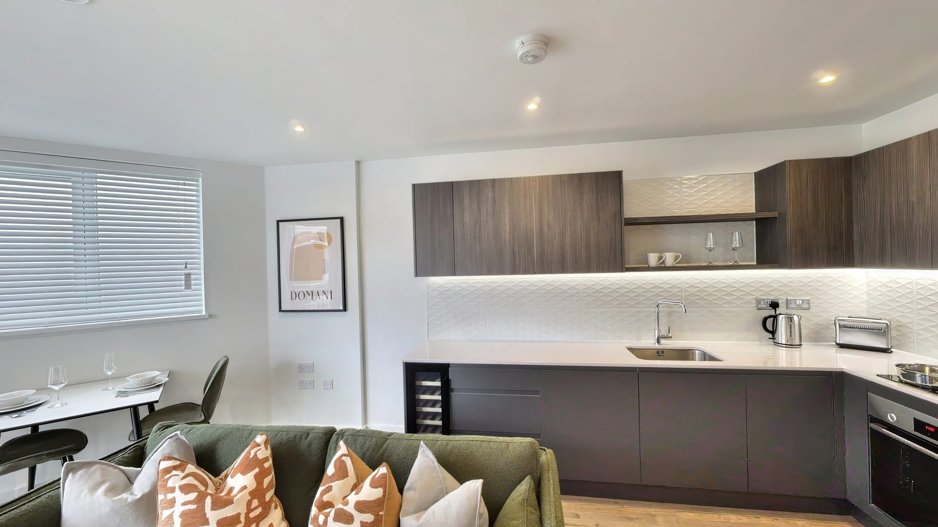 Living room in Rosa House with a green sofa and terracotta cushions, opening onto a walnut kitchen